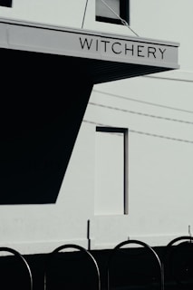 A modern building facade with a sign that reads 'Witchery' is prominently displayed. The structure casts a large shadow against a light-colored wall, beneath which lies a row of curved metal bars or bicycle racks. The image captures a clear day with visible shadows due to the sun.