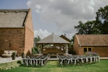 An outdoor event setup with rows of chairs adorned with white fabric, positioned on a green lawn facing a small wooden pavilion. The pavilion is decorated with hanging white paper lanterns and surrounded by brick buildings, under a cloudy sky.