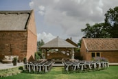 An outdoor event setup with rows of chairs adorned with white fabric, positioned on a green lawn facing a small wooden pavilion. The pavilion is decorated with hanging white paper lanterns and surrounded by brick buildings, under a cloudy sky.