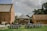 An outdoor event setup with rows of chairs adorned with white fabric, positioned on a green lawn facing a small wooden pavilion. The pavilion is decorated with hanging white paper lanterns and surrounded by brick buildings, under a cloudy sky.