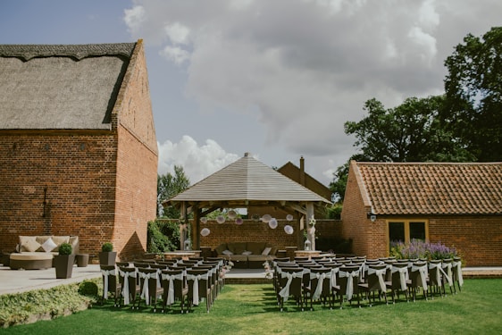 An outdoor event setup with rows of chairs adorned with white fabric, positioned on a green lawn facing a small wooden pavilion. The pavilion is decorated with hanging white paper lanterns and surrounded by brick buildings, under a cloudy sky.