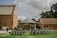 An outdoor event setup with rows of chairs adorned with white fabric, positioned on a green lawn facing a small wooden pavilion. The pavilion is decorated with hanging white paper lanterns and surrounded by brick buildings, under a cloudy sky.