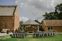 An outdoor event setup with rows of chairs adorned with white fabric, positioned on a green lawn facing a small wooden pavilion. The pavilion is decorated with hanging white paper lanterns and surrounded by brick buildings, under a cloudy sky.