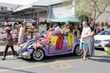 A street market scene where a purple vintage car is covered with various clothing items hanging from it. People are browsing and selling goods, including a man walking past and a man and a woman standing by the car. There are market stalls set up with hats and more clothing items, and a building with a corrugated metal roof and barbed wire fence in the background. The market appears busy and lively with diverse merchandise on display.