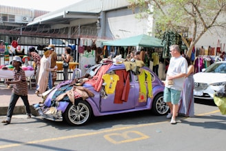 A street market scene where a purple vintage car is covered with various clothing items hanging from it. People are browsing and selling goods, including a man walking past and a man and a woman standing by the car. There are market stalls set up with hats and more clothing items, and a building with a corrugated metal roof and barbed wire fence in the background. The market appears busy and lively with diverse merchandise on display.