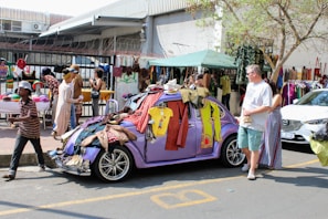 A street market scene where a purple vintage car is covered with various clothing items hanging from it. People are browsing and selling goods, including a man walking past and a man and a woman standing by the car. There are market stalls set up with hats and more clothing items, and a building with a corrugated metal roof and barbed wire fence in the background. The market appears busy and lively with diverse merchandise on display.