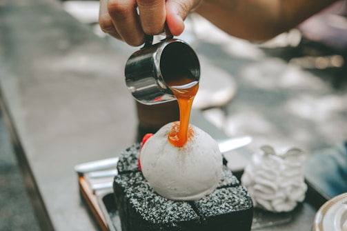 A hand is pouring a caramel sauce from a metal jug onto a spherical dessert. The dessert is sitting atop a square platform of what seems to be chocolate cake dusted with powdered sugar. To the side, there is a textured, cream-like decoration.