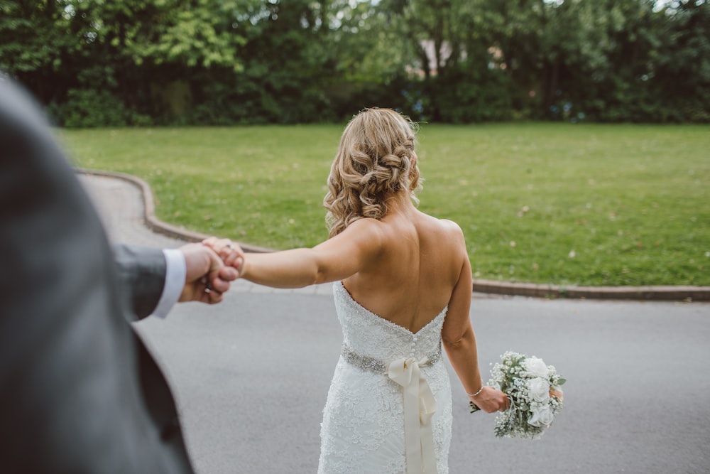 Braut im rückenfreien Brautkleid hält Blumenstrauß, während sie die Hand des Bräutigams von hinten hält
