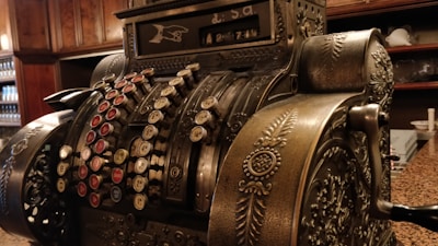 An ornate vintage cash register with intricate metalwork and a series of colorful round keys. The machine features elaborate decorative patterns and a mechanical display at the top. It's set against a background of wooden shelves and a countertop.