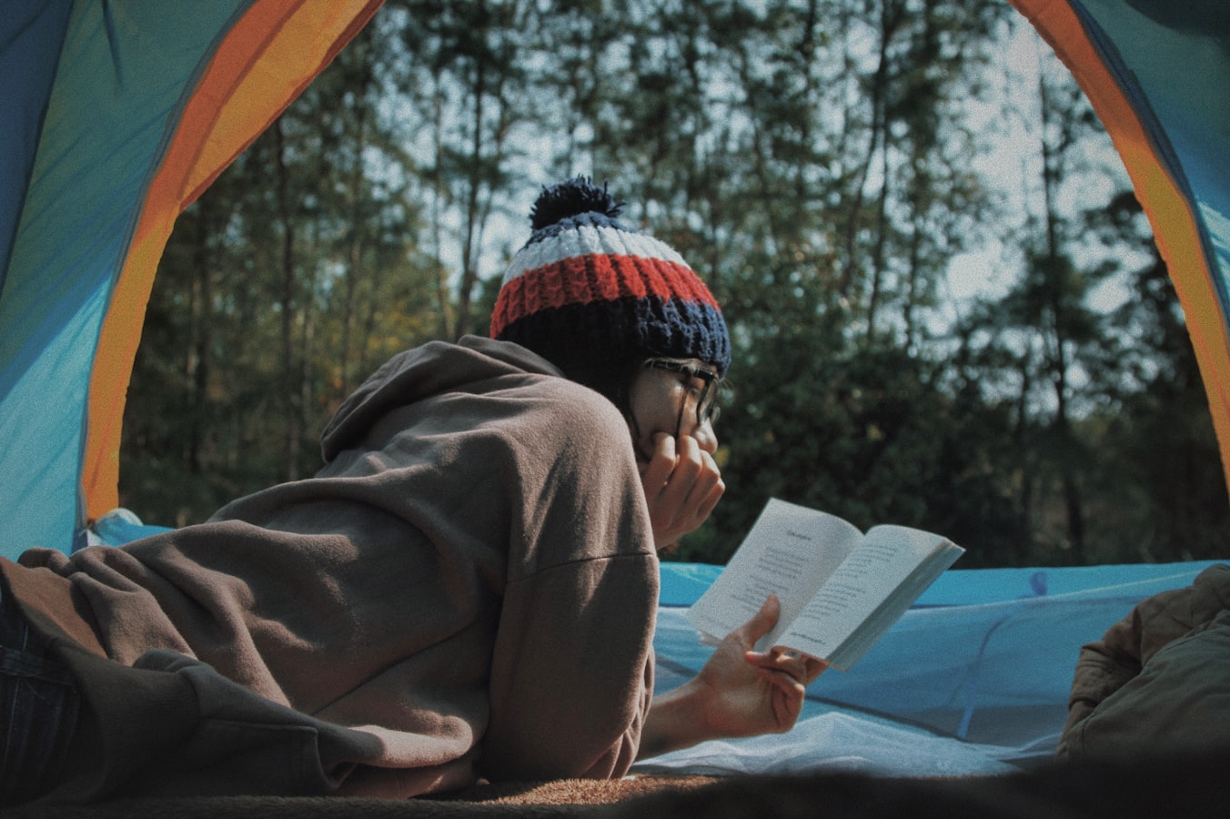 A young woman with a red, white, and blue beanie and glasses reading a book.