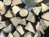 A stack of chopped wooden logs arranged in a neat pile. The logs have a visible, freshly cut appearance with light brown hues and distinct growth rings and textures.