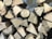 A stack of chopped wooden logs arranged in a neat pile. The logs have a visible, freshly cut appearance with light brown hues and distinct growth rings and textures.