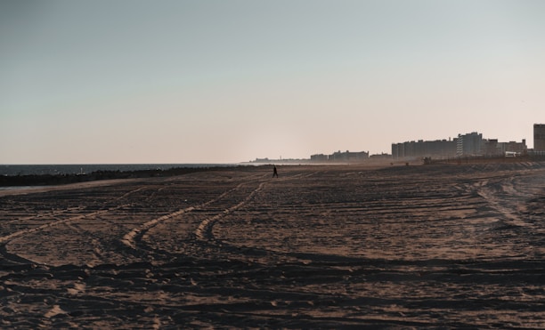A sandy beach extends into the distance with visible tire tracks running across it. The ocean lies to the left, with waves gently lapping against the shore. On the right, there are several tall buildings, likely apartments or hotels, lining the horizon. A lone figure is walking along the beach, adding a sense of solitude.