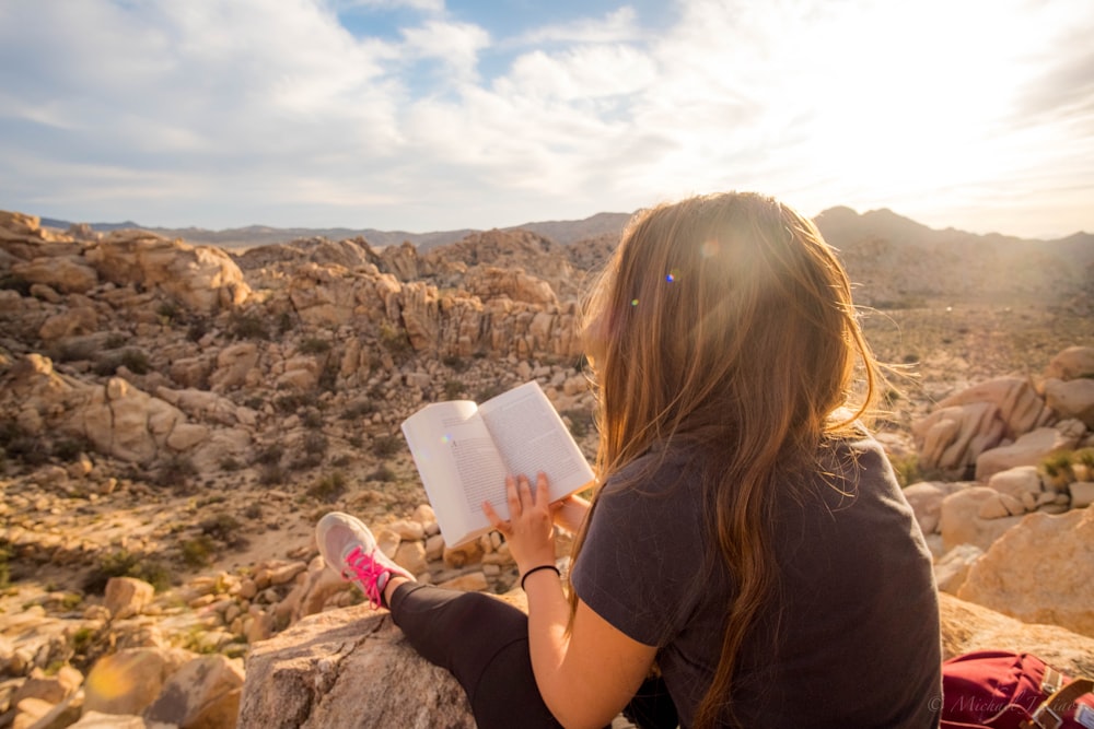 woman sitting on rock formation while reading book