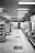 A black and white image depicts an empty aisle in a grocery store. Shelves line both sides, stocked with various products. Bright overhead lighting reflects off the clean, checkered floor tiles.