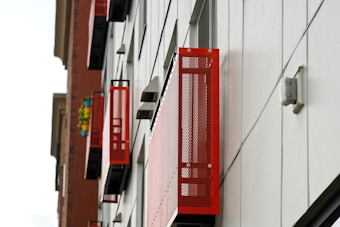 A series of red metal balconies are attached to the side of a modern building with white paneling. The building has a sleek and industrial appearance, and the red elements stand out vividly against the neutral background. A brick building is visible in the background, featuring colorful potted plants on a balcony.