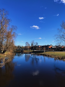 A serene river scene with clear blue skies and a few scattered clouds. The river reflects the sky and nearby trees, creating a mirror-like surface. Bare trees line the riverbank, and a distant bridge is visible along with modern buildings.