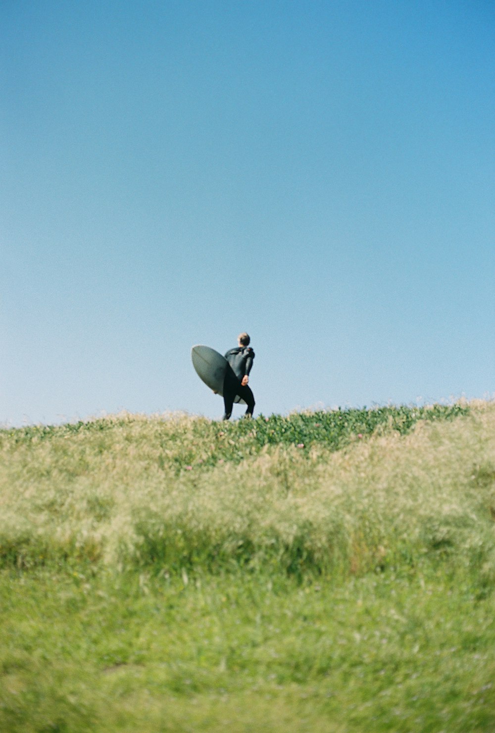 man standing on green grass mountain holding long surfboard