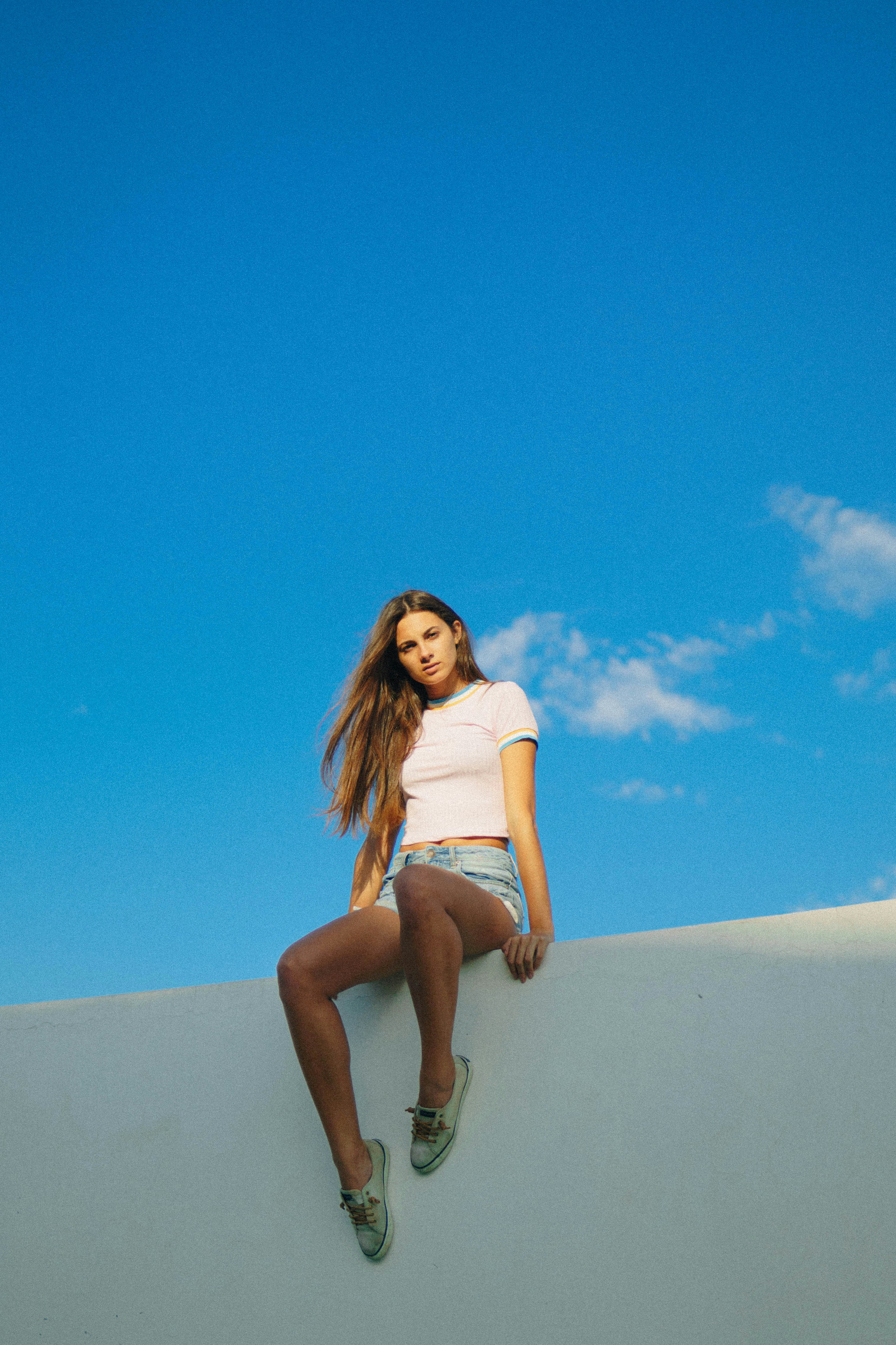 woman wearing white crop top and blue denim shorts while sitting on top of white wall during clear blue sky