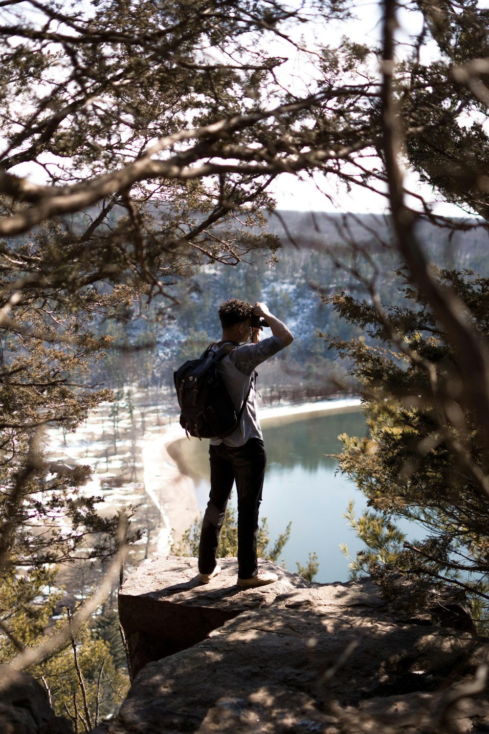 회색 드레스 셔츠와 검은 가방을 입은 남자가 바위 위에 서서 호수 앞에서 사진을 찍고 있습니다.