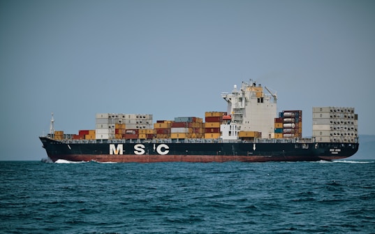 A large cargo ship is sailing on the ocean, loaded with numerous colorful shipping containers stacked on its deck. The ship has a black hull and white superstructure, with the letters 'MSC' prominently displayed on the side. The background is a clear horizon with a slightly overcast sky.