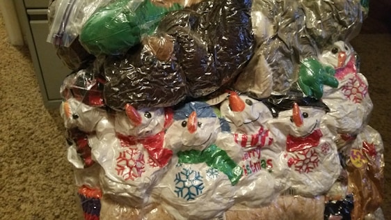 A collection of plastic bags, many featuring snowmen with red scarves and orange carrot noses, is tightly packed together. The bags are colorful, with various designs including snowflakes and holiday greetings. The stuffed formation has a prominent texture due to the crumpling and layering of the plastic.