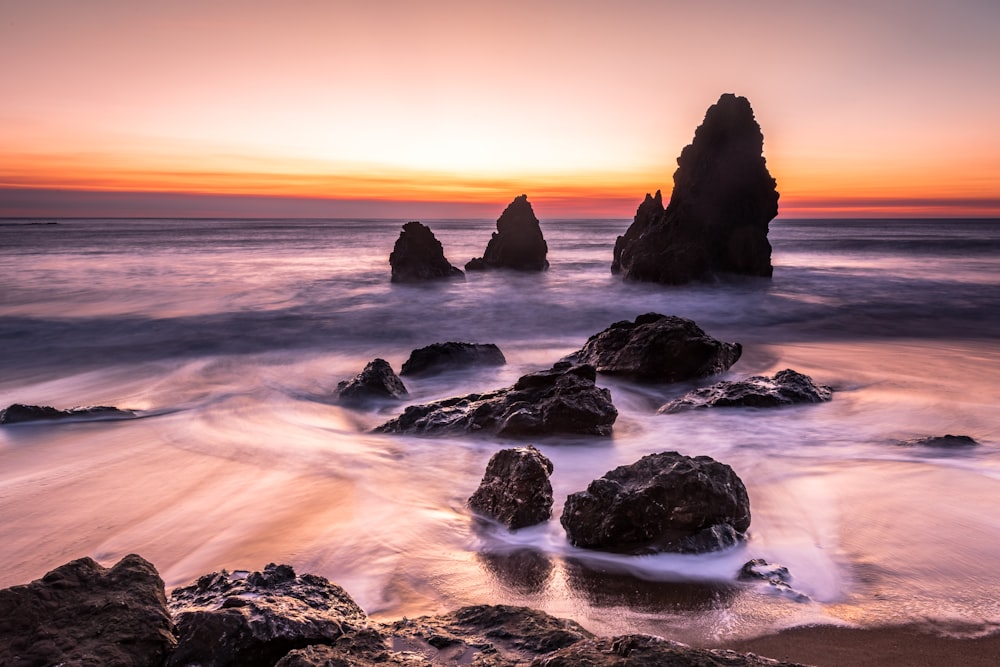 Fotografie des Strandes in der Nähe einer schwarzen Felsformation unter orangefarbenem Himmel