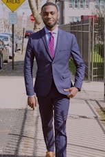 man in blue suit walking on the sidewalk near metal fence and parked vehicle