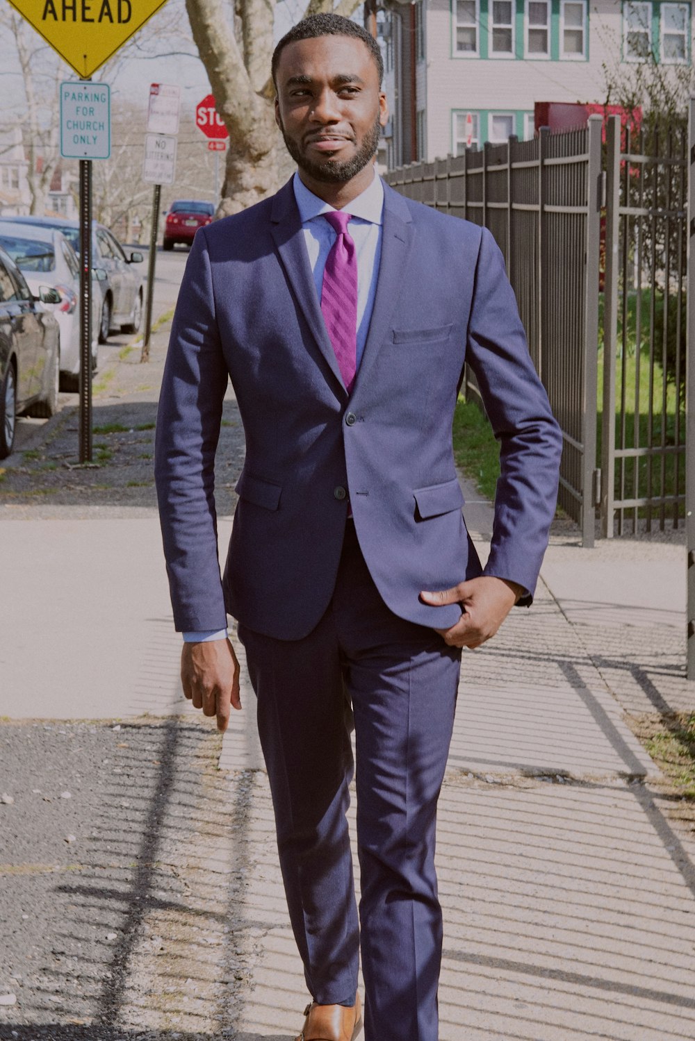 man in blue suit walking on the sidewalk near metal fence and parked vehicle