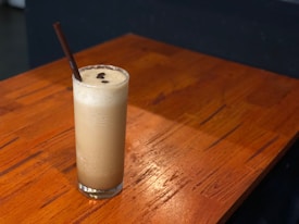 A tall glass filled with a frothy, creamy beverage sits on a wooden table. The drink has a light brown color and is topped with a few coffee beans. A chocolate-colored straw is placed in the glass, and the wooden surface has a warm, polished finish with visible wood grain patterns.