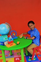 A young child is sitting at a small, green table on a red chair, playing with toy vehicles. The table has a globe, a colorful clock, blocks, and a small figure. The floor is covered with colorful lettered play mats, and the background is a bright orange wall.