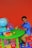 A young child is sitting at a small, green table on a red chair, playing with toy vehicles. The table has a globe, a colorful clock, blocks, and a small figure. The floor is covered with colorful lettered play mats, and the background is a bright orange wall.