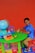 A young child is sitting at a small, green table on a red chair, playing with toy vehicles. The table has a globe, a colorful clock, blocks, and a small figure. The floor is covered with colorful lettered play mats, and the background is a bright orange wall.