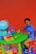 A young child is sitting at a small, green table on a red chair, playing with toy vehicles. The table has a globe, a colorful clock, blocks, and a small figure. The floor is covered with colorful lettered play mats, and the background is a bright orange wall.