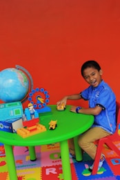 A young child is sitting at a small, green table on a red chair, playing with toy vehicles. The table has a globe, a colorful clock, blocks, and a small figure. The floor is covered with colorful lettered play mats, and the background is a bright orange wall.