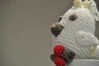 A handmade crocheted toy with various colors, primarily white, with brown and red accents. The plush has a rounded shape, with small crocheted elements on its surface, possibly representing facial features or decorations. It appears to be sitting inside or on the edge of a white ceramic cup. The background is plain and neutral, allowing the toy's details to stand out.