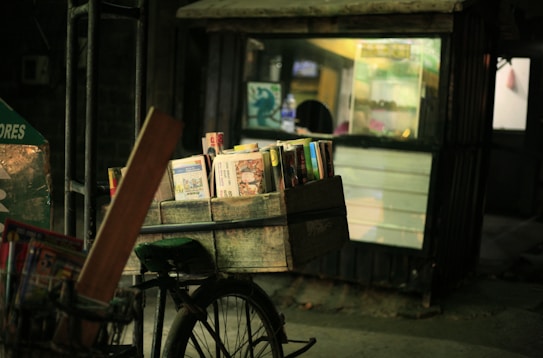 A bicycle with a wooden crate carrying books is parked in front of a small shop. The shop has a partially illuminated display and appears to be closed, with some lights on inside. The setting is dimly lit, giving a quiet, nighttime street scene vibe.