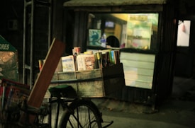 A bicycle with a wooden crate carrying books is parked in front of a small shop. The shop has a partially illuminated display and appears to be closed, with some lights on inside. The setting is dimly lit, giving a quiet, nighttime street scene vibe.