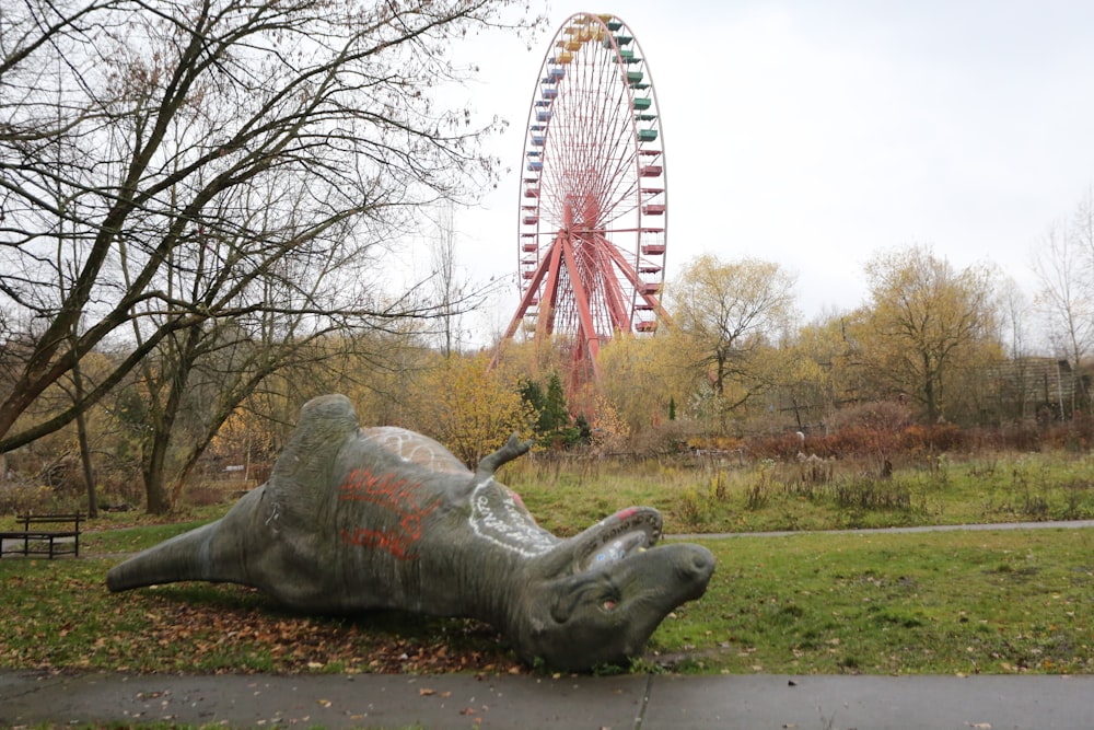 estatua de dinosaurio de hormigón gris que yace en la hierba verde cerca de la rueda de la fortuna roja