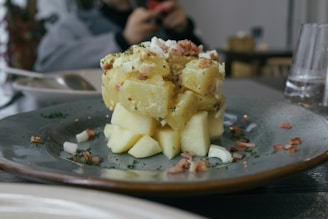 A plate with a carefully arranged potato salad topped with chopped onions and herbs. Bacon bits are scattered around, and the salad is served on a gray ceramic plate. There are blurred objects in the background, indicating a dining setting.