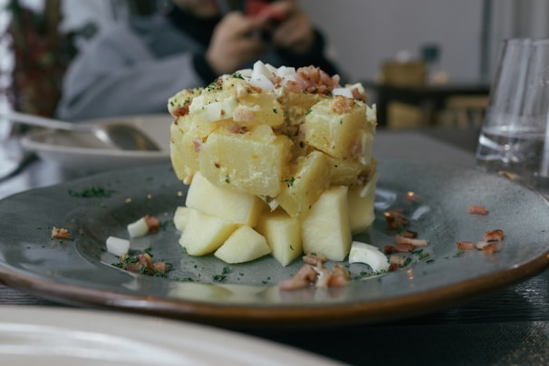 A plate with a carefully arranged potato salad topped with chopped onions and herbs. Bacon bits are scattered around, and the salad is served on a gray ceramic plate. There are blurred objects in the background, indicating a dining setting.