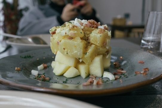 A plate with a carefully arranged potato salad topped with chopped onions and herbs. Bacon bits are scattered around, and the salad is served on a gray ceramic plate. There are blurred objects in the background, indicating a dining setting.