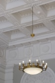 An ornate ceiling features intricate plasterwork and a decorative pattern of flowers and geometric shapes. Hanging from the center is an elegant chandelier with multiple small light bulbs on a circular frame, made from metal with an antique finish. The walls complement the ceiling with ornate embellishments in a similar style.