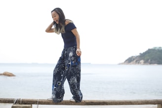 A woman with long hair laughs joyfully while balancing on a wooden beam in front of a serene beach backdrop. She wears a dark top and loose, patterned pants with white stars and designs. The sky is overcast, and the sea is calm.