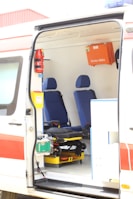 The interior of an ambulance is visible with two blue seats and various medical equipment and storage compartments. A first aid box labeled 'Erste Hilfe' is mounted on the wall. The van's door is open, showing a stretcher and other emergency tools.