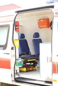 The interior of an ambulance is visible with two blue seats and various medical equipment and storage compartments. A first aid box labeled 'Erste Hilfe' is mounted on the wall. The van's door is open, showing a stretcher and other emergency tools.