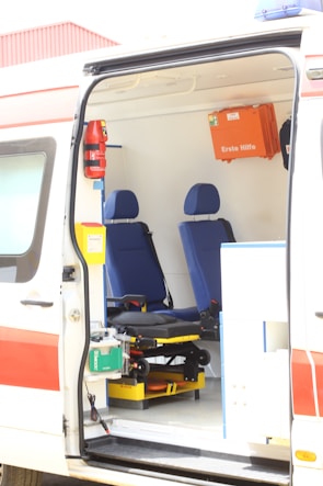 The interior of an ambulance is visible with two blue seats and various medical equipment and storage compartments. A first aid box labeled 'Erste Hilfe' is mounted on the wall. The van's door is open, showing a stretcher and other emergency tools.