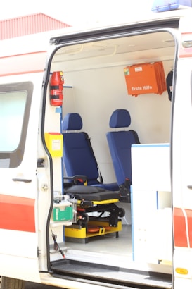 The interior of an ambulance is visible with two blue seats and various medical equipment and storage compartments. A first aid box labeled 'Erste Hilfe' is mounted on the wall. The van's door is open, showing a stretcher and other emergency tools.