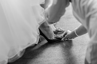 A close-up in black and white of a person helping someone else put on a high-heeled shoe adorned with decorative embellishments. The scene captures a moment of intimate assistance, likely involving formal attire such as a gown or a dress.