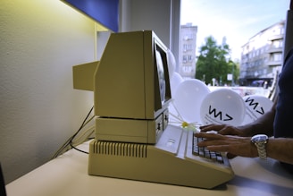 A person is typing on an old beige computer, possibly from the 1980s, situated in an office setting near a window. Outside, a cityscape with trees and modern buildings is visible. White balloons with printed logos are floating beside the computer.